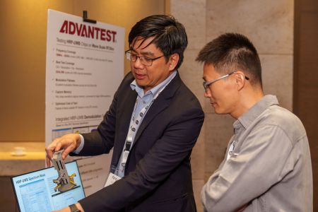 Two men at a tech booth review data on a laptop while one holds an electronic device.